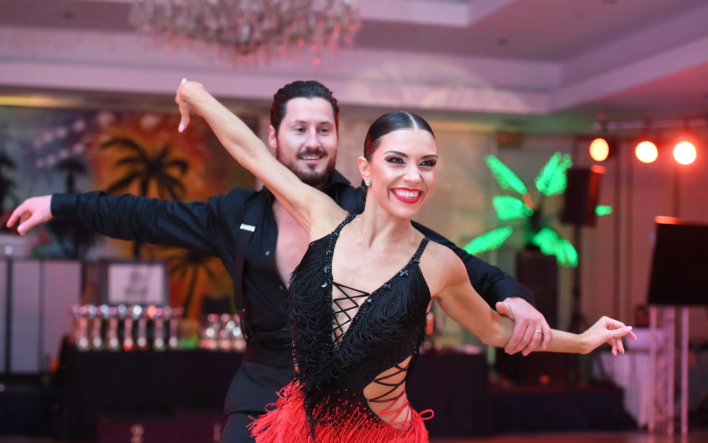 A smiling Latin dance couple mid-turn on the floor at a Dance With Me showcase